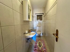 Een kleine badkamer met witte tegels, een wastafel, toilet en een raam met zonwering.