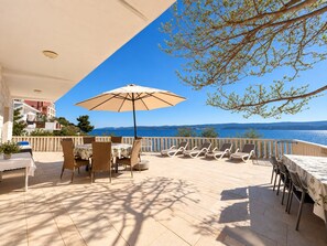 An outdoor terrace with a table and chairs, a large umbrella, and a view of the sea.