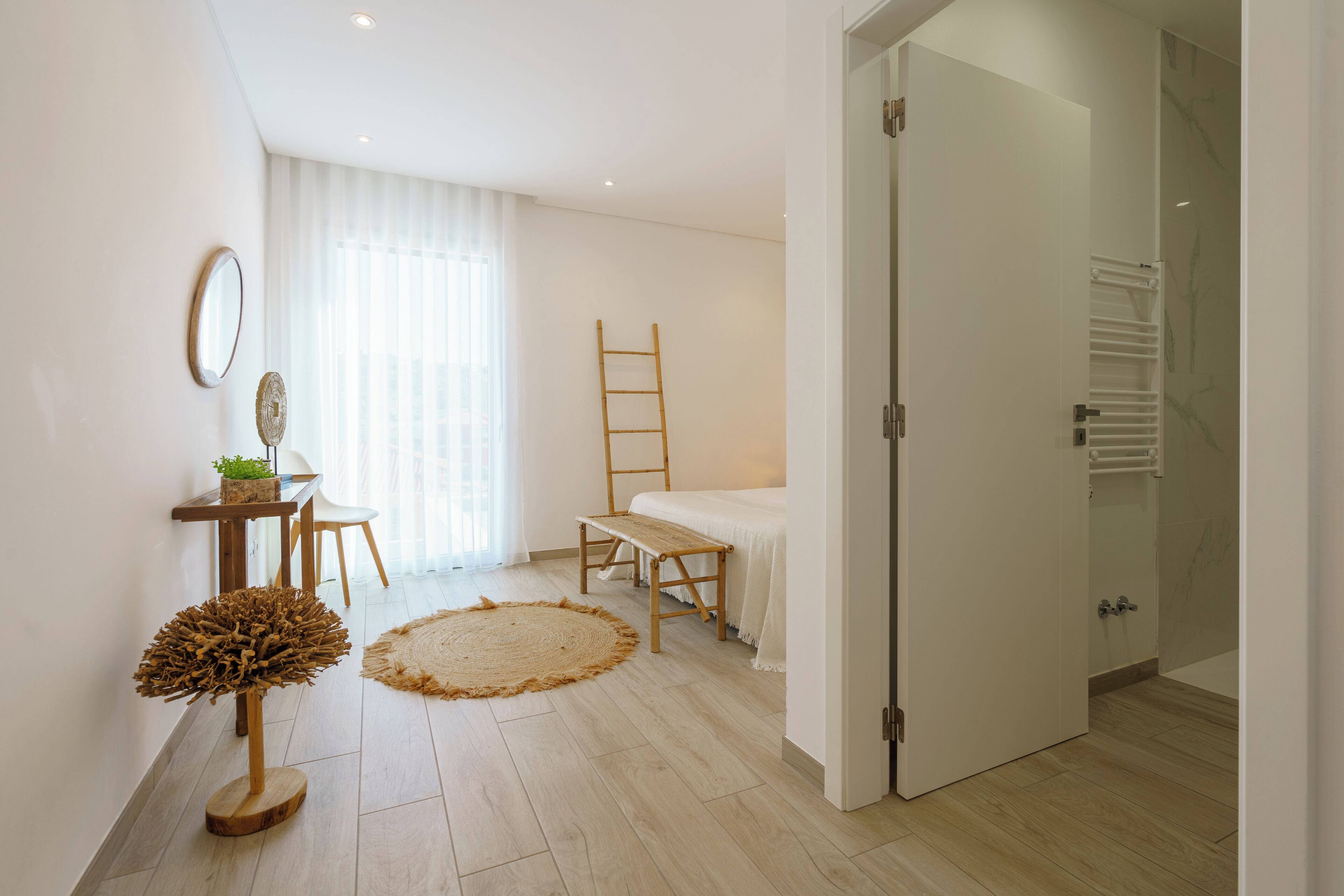 Een moderne slaapkamer met een bed, een houten trap, een bureau met een plant en een groot raam met dunne gordijnen.