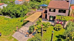 Ein zweigeschossiges Haus mit Ziegeldach, überdachter Terrasse und Garten mit Palmen.