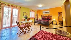 Ein Zimmer mit einem Bett, einem Esstisch mit Stühlen, einem kleinen Tisch mit einer Vase und einem Fenster mit Vorhängen.