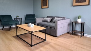 Een moderne woonkamer met een grijze bank, twee donkergroene fauteuils, een houten salontafel en twee bijzettafeltjes met planten.