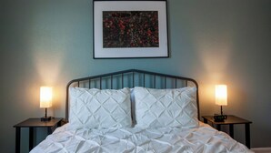 Een netjes opgemaakt bed met wit beddengoed, twee nachtkastjes met lampen, en een ingelijst kunstwerk aan de muur.