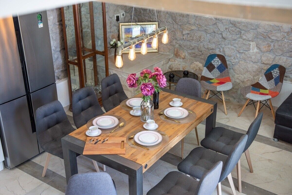 Un coin repas avec une table en bois dressée pour six personnes, entourée de chaises modernes. On aperçoit un mur en pierre et une cheminée en pierre à l’arrière-plan.