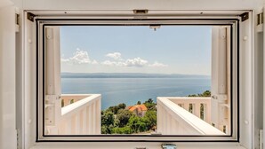 Une fenêtre donnant sur un paysage côtier, avec une étendue d’eau et des montagnes à l’horizon.