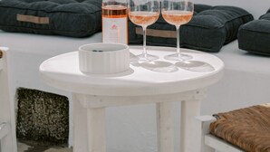 Una mesa blanca con una botella y dos vasos de líquido rosado, un cuenco blanco y cojines oscuros.