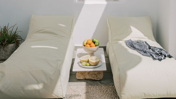 Una habitación iluminada por el sol con un ventanal pequeño, dos sofás color beige, una bandeja con frutas y una planta.