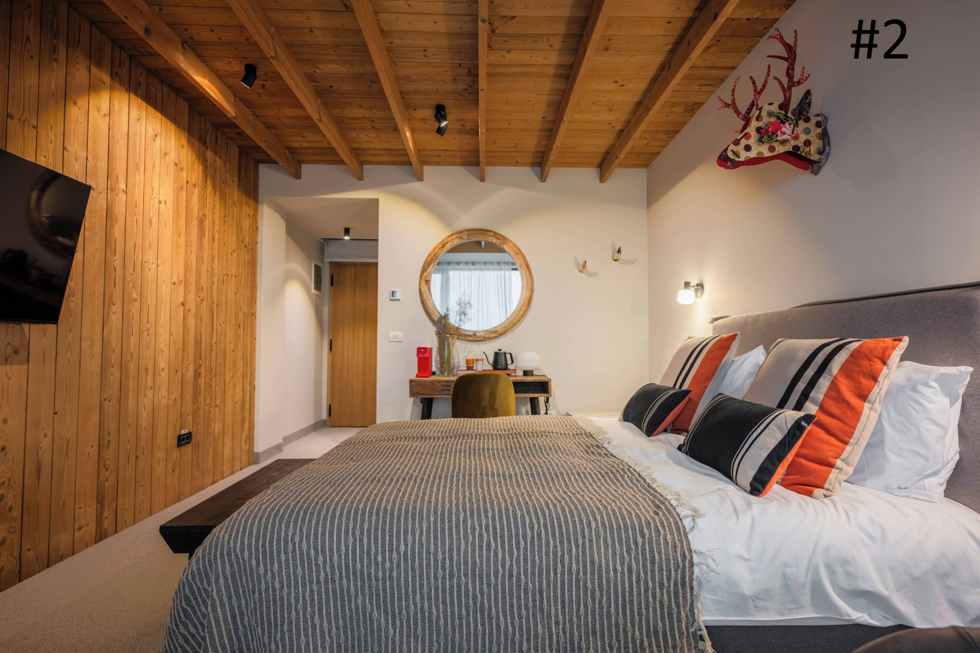 A bedroom with a wooden ceiling, a bed with striped bedding, a round mirror, and a wall-mounted TV.