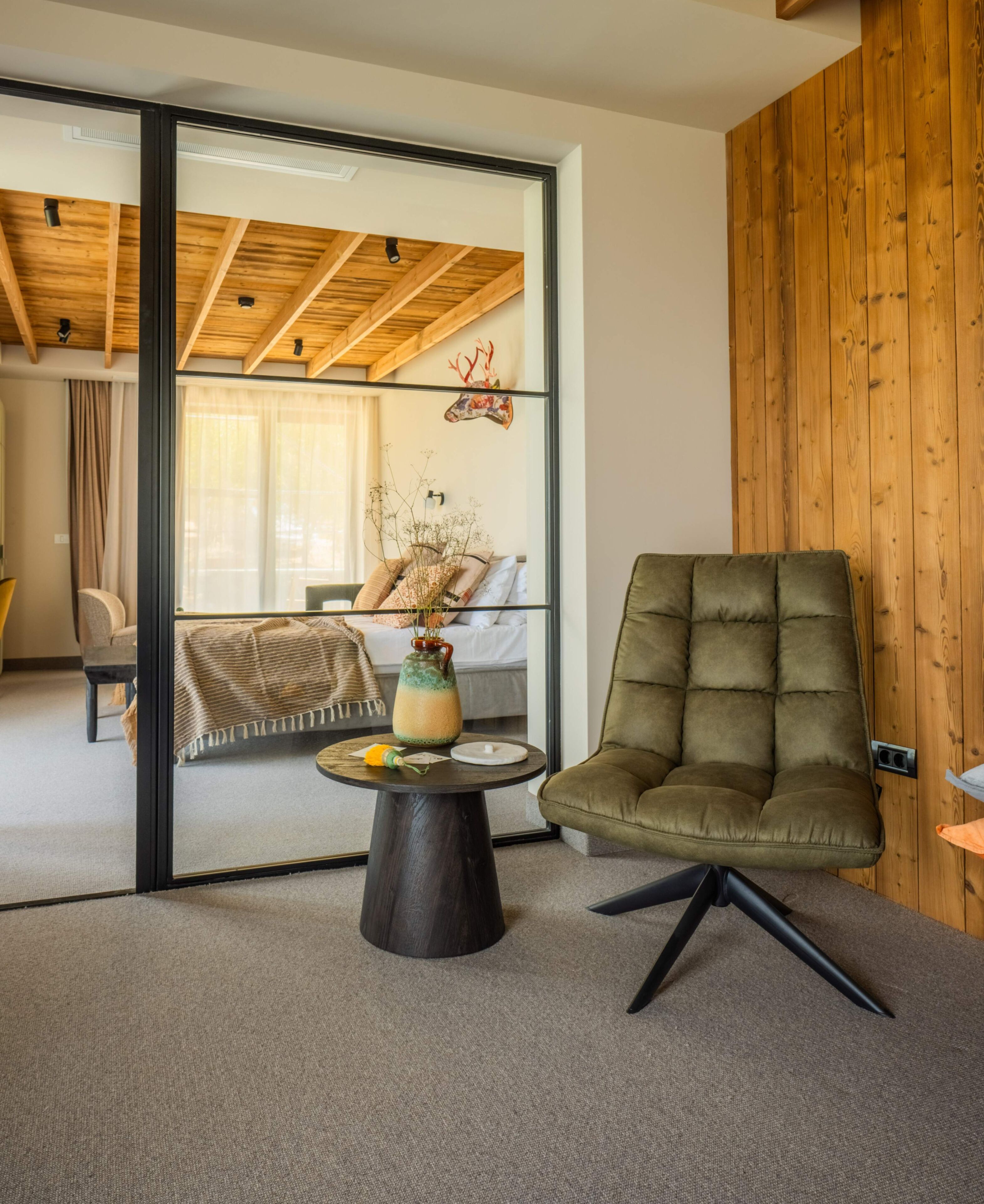 Una sala de estar moderna con un ventanal de vidrio que da a otra habitación, un sillón verde, una mesa de centro redonda con un florero y un techo de madera.