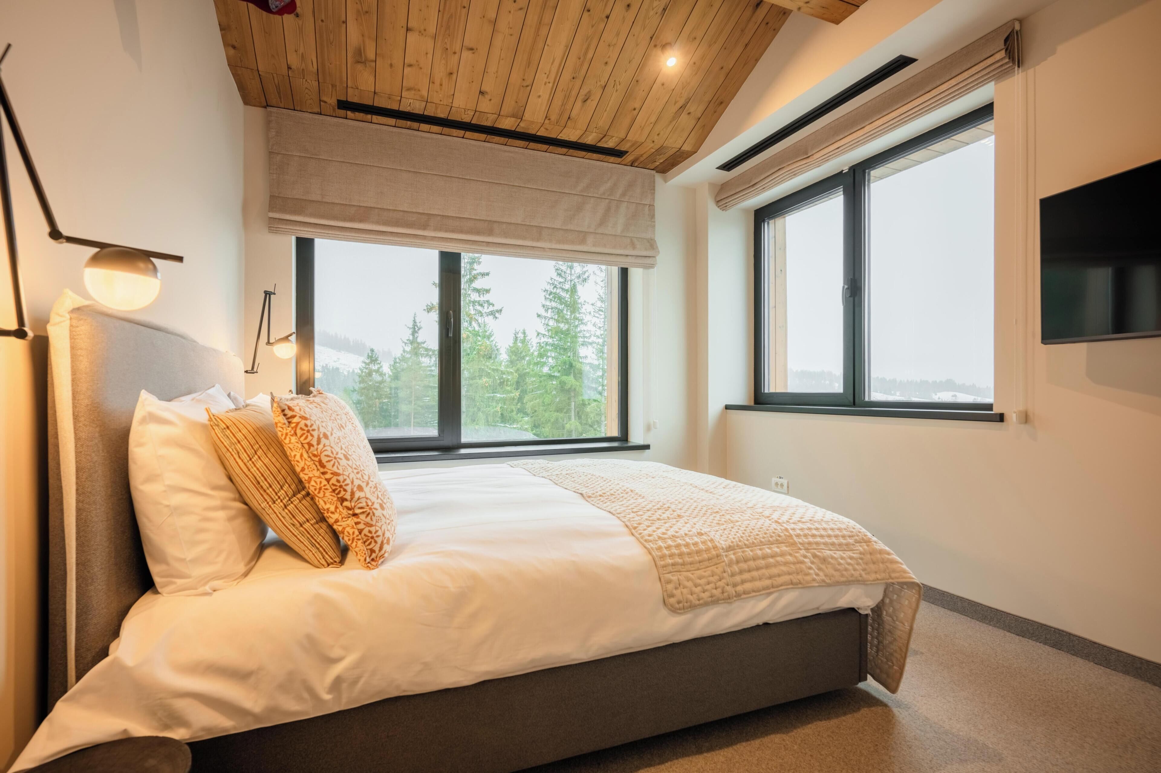 A bedroom with a large bed, two windows, and a view of trees.