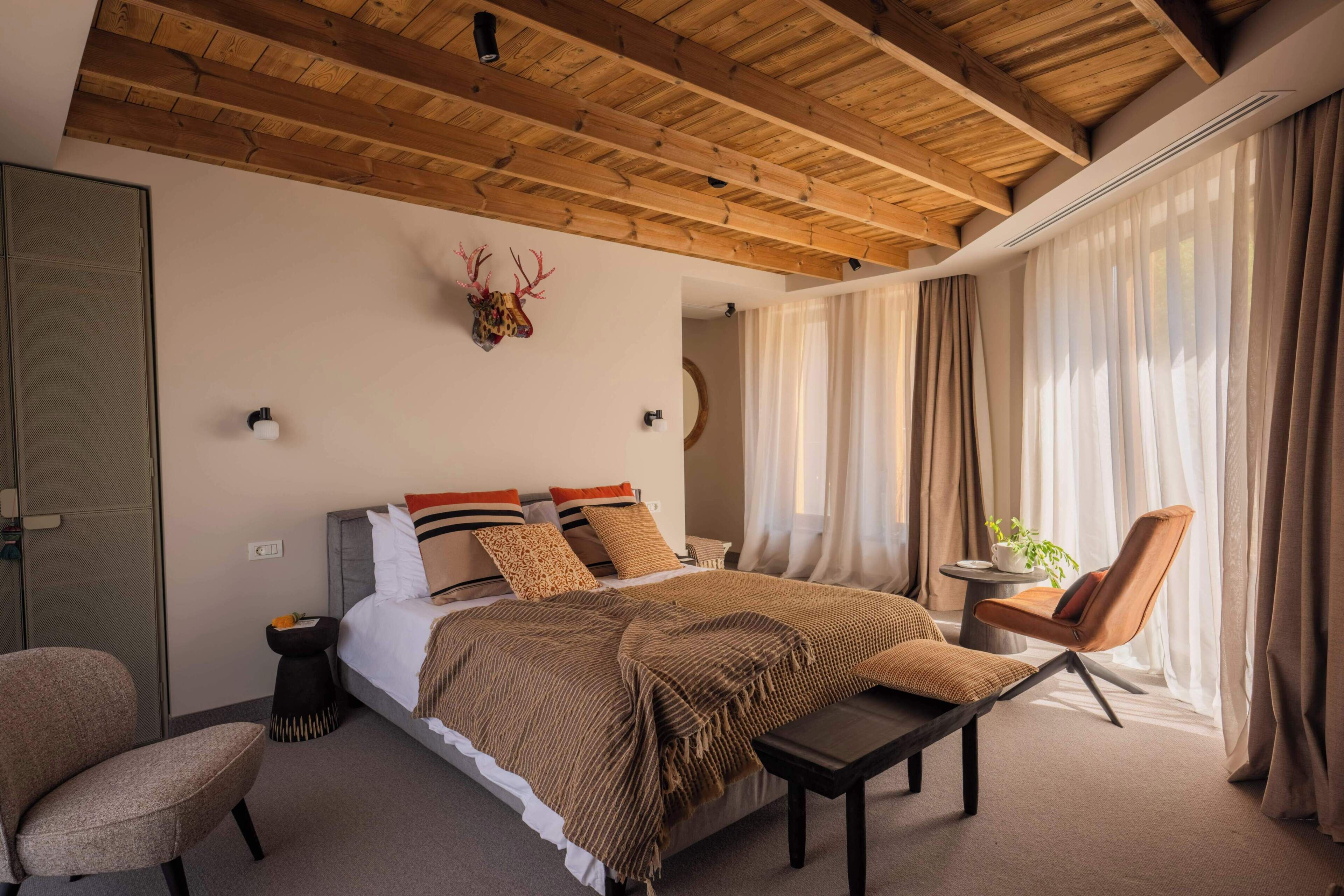 A bedroom with a wooden ceiling, a bed with a brown blanket, a chair, and a small table with a plant.