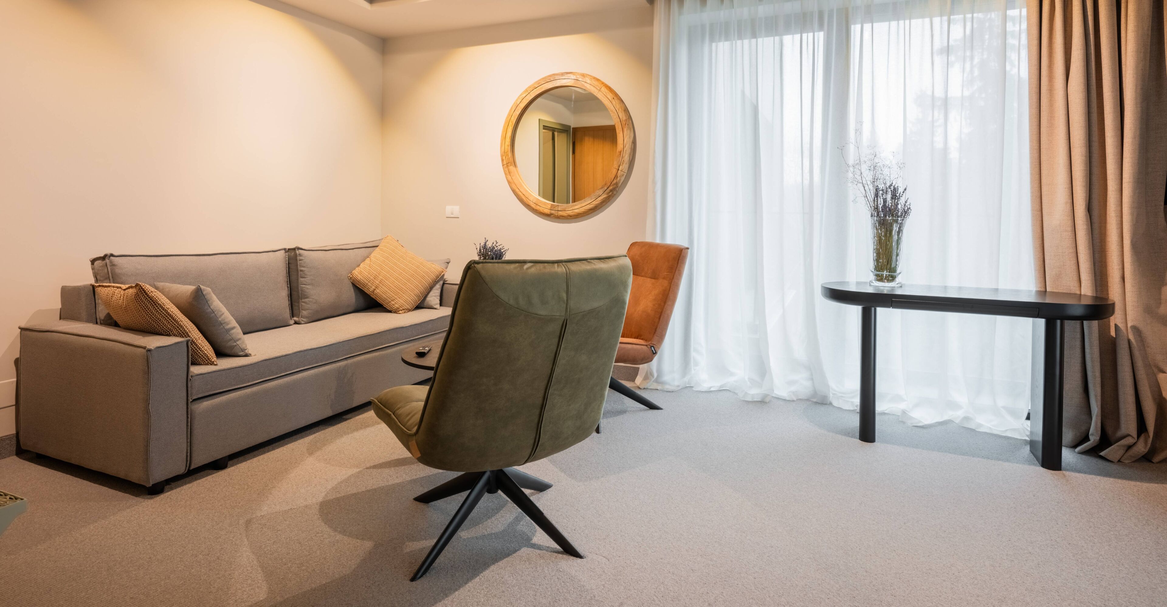 A modern living room with a grey sofa, a green armchair, a round mirror, and a small table with a vase.