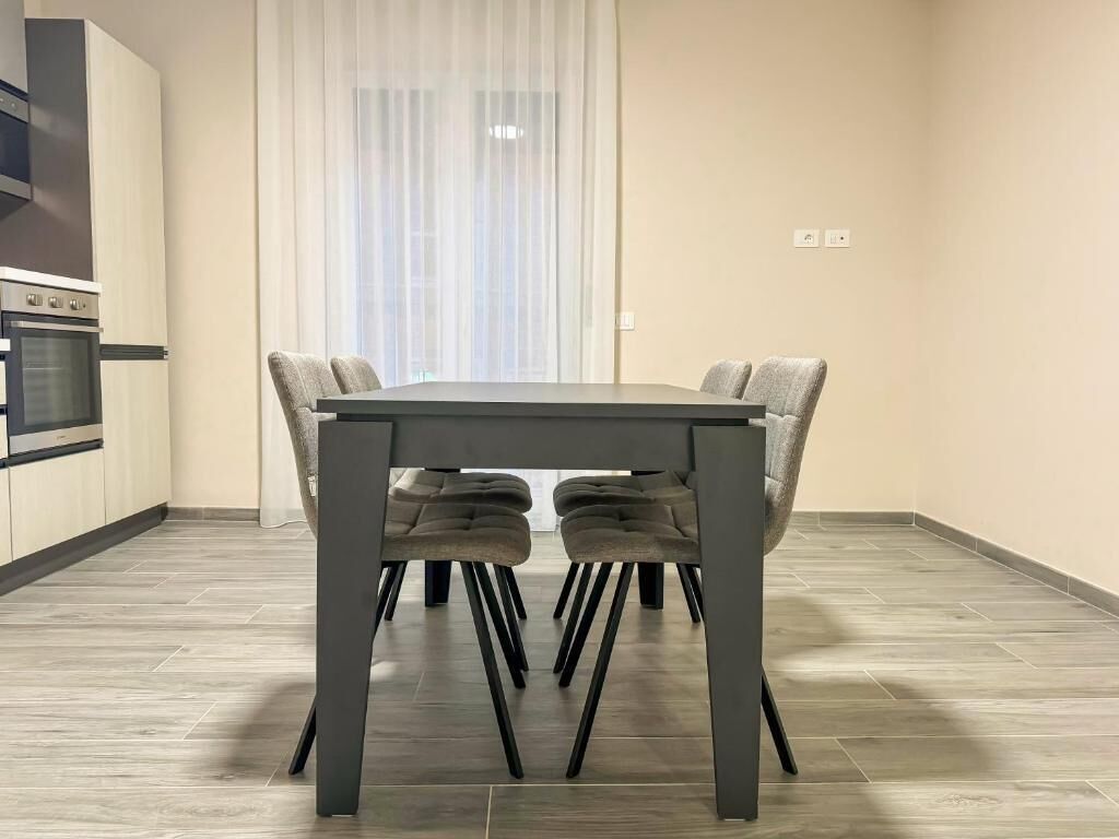 A modern kitchen with a dining table and chairs, a built-in oven, and a window with sheer curtains.
