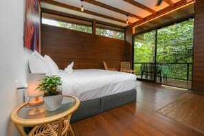 Un dormitorio con una cama grande, una mesita de noche con una lámpara y una planta, una ventana con vista a zonas verdes y un balcón con sillas.