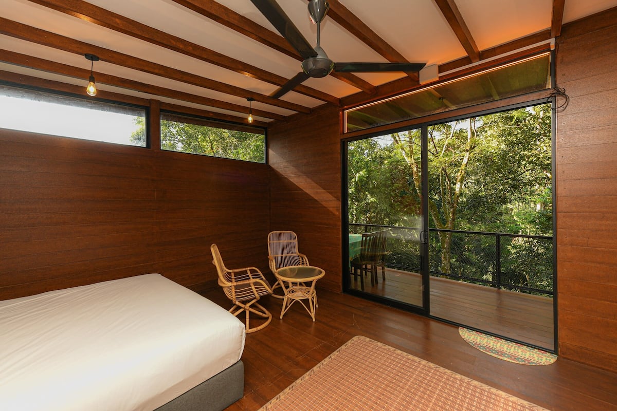 Quarto com cama, duas cadeiras de vime, uma mesinha e varanda com vista para árvores.