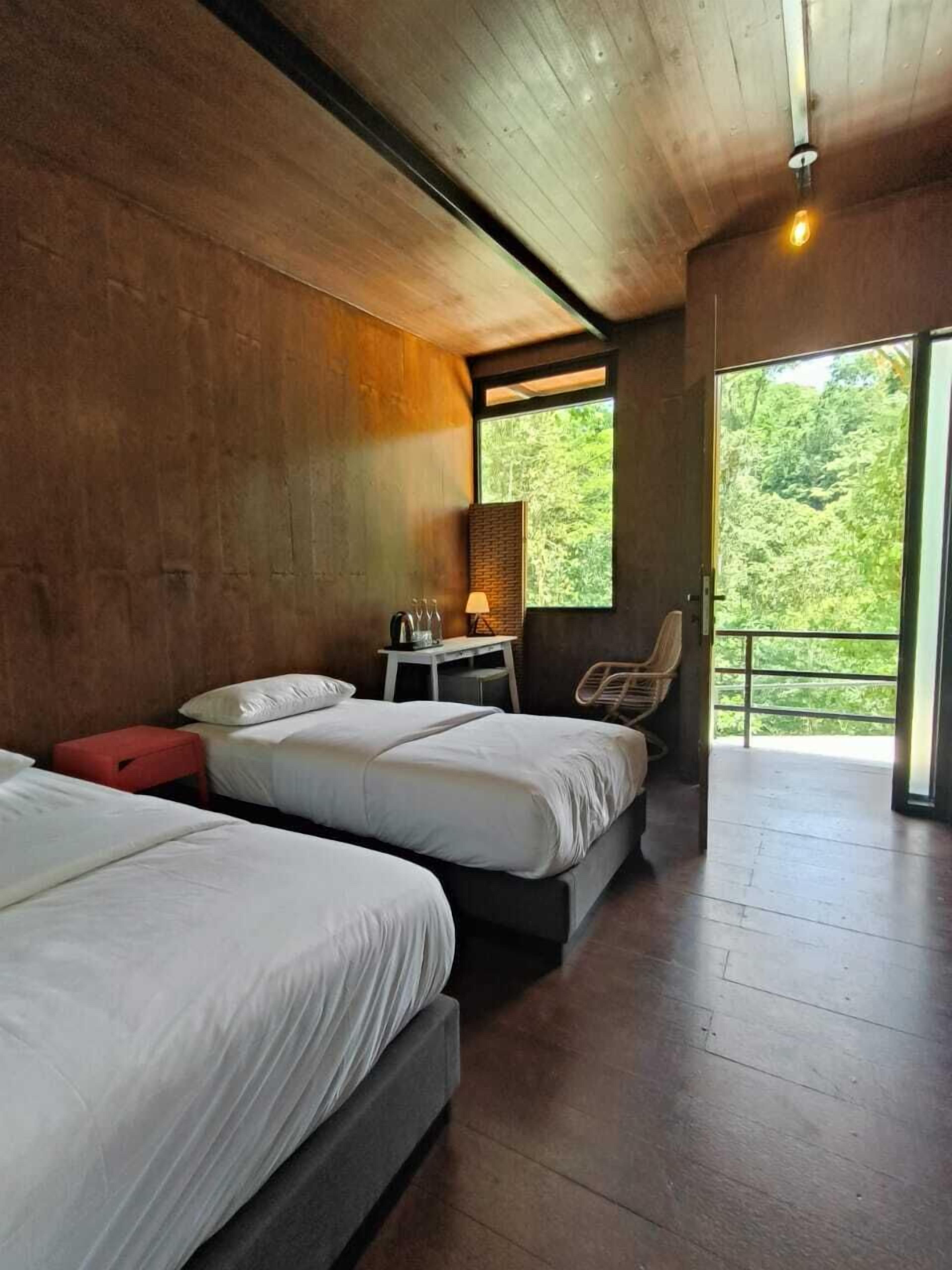 A room with two beds, a small table with a lamp, a chair, and a large window with a view of trees.