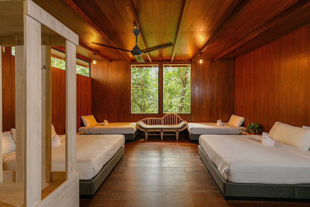 Quarto com duas camas, um banco e amplas janelas com vista para árvores.