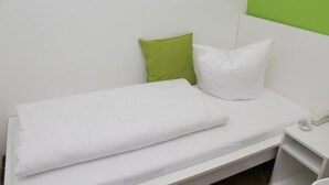 Ein kleines Zimmer mit einem Einzelbett, weißer Bettwäsche und einem grünen Kopfkissen.