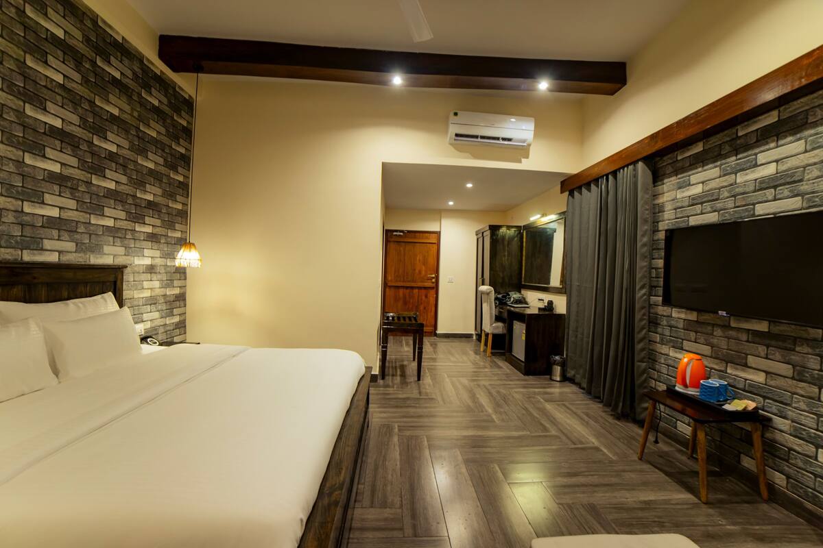 Quarto de hotel com uma cama grande, uma escrivaninha com cadeira, uma televisão na parede e parede com padrão de tijolos.