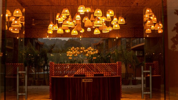 A reception area with a wooden reception desk, multiple hanging lamps, and a view of outdoor greenery. - Vanya Forest Resort (Kaladhungi)