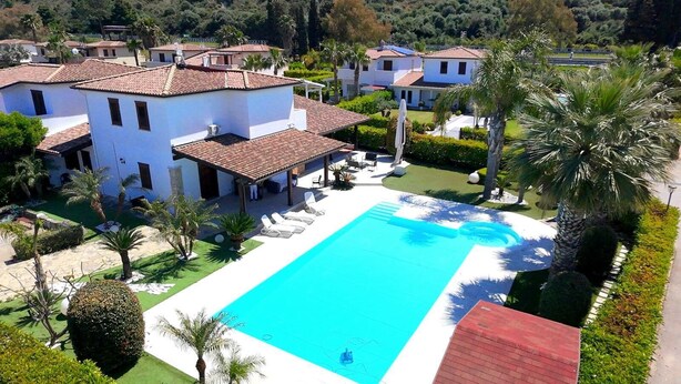 A large swimming pool surrounded by a well-maintained garden and a white building with a red roof.