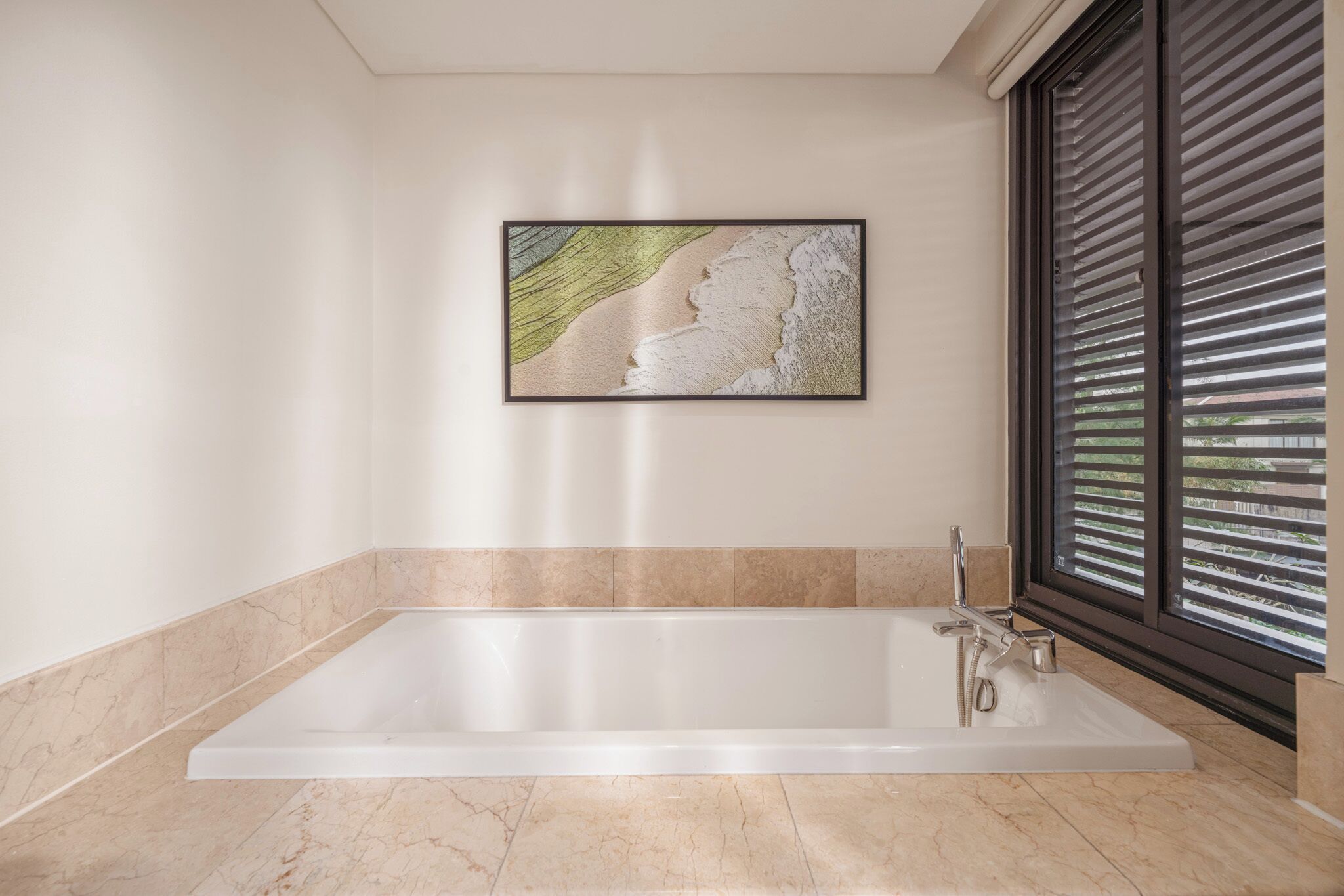 A modern bathroom with a large bathtub, a framed artwork on the wall, and a window with blinds.