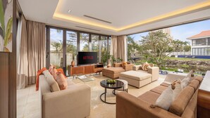 A modern living room with a large TV, comfortable seating, and a view of a garden and building. - Ocean Villa Da Nang 4BR 5 Beds, Private Pool and Kitchen (Da Nang)