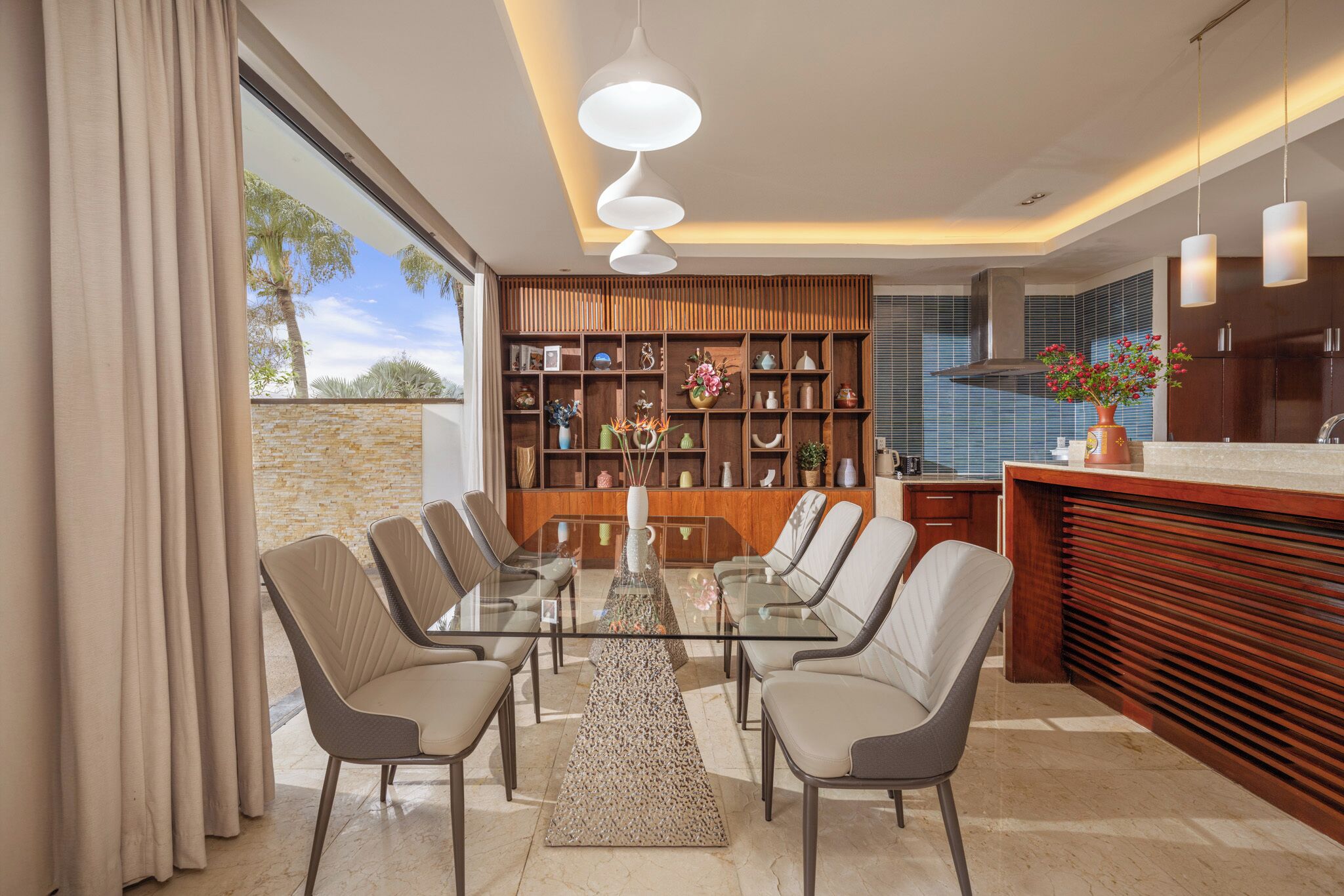 A modern kitchen with a glass dining table, wooden shelves, and pendant lighting.