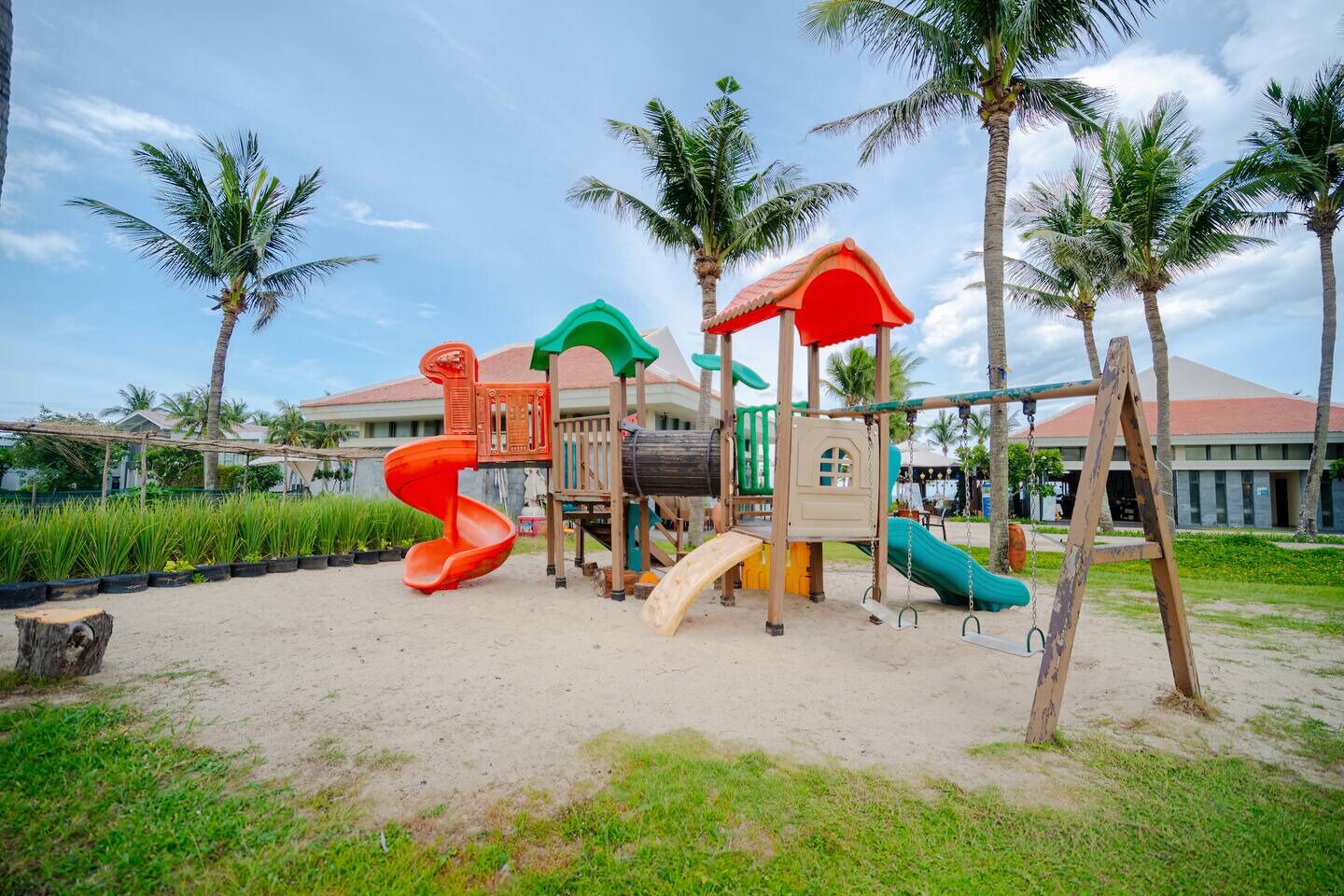 A playground with slides and climbing structures, surrounded by palm trees and a building in the background.