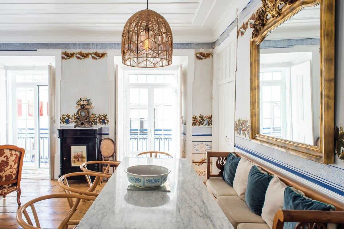 Un coin repas comprenant une table en marbre, des chaises en osier, un canapé avec des coussins, un miroir décoratif et un luminaire suspendu.