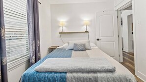 Ein Schlafzimmer mit einem Bett, zwei Nachttischlampen, einem Fenster mit Jalousien und einer Tür zu einem anderen Raum.