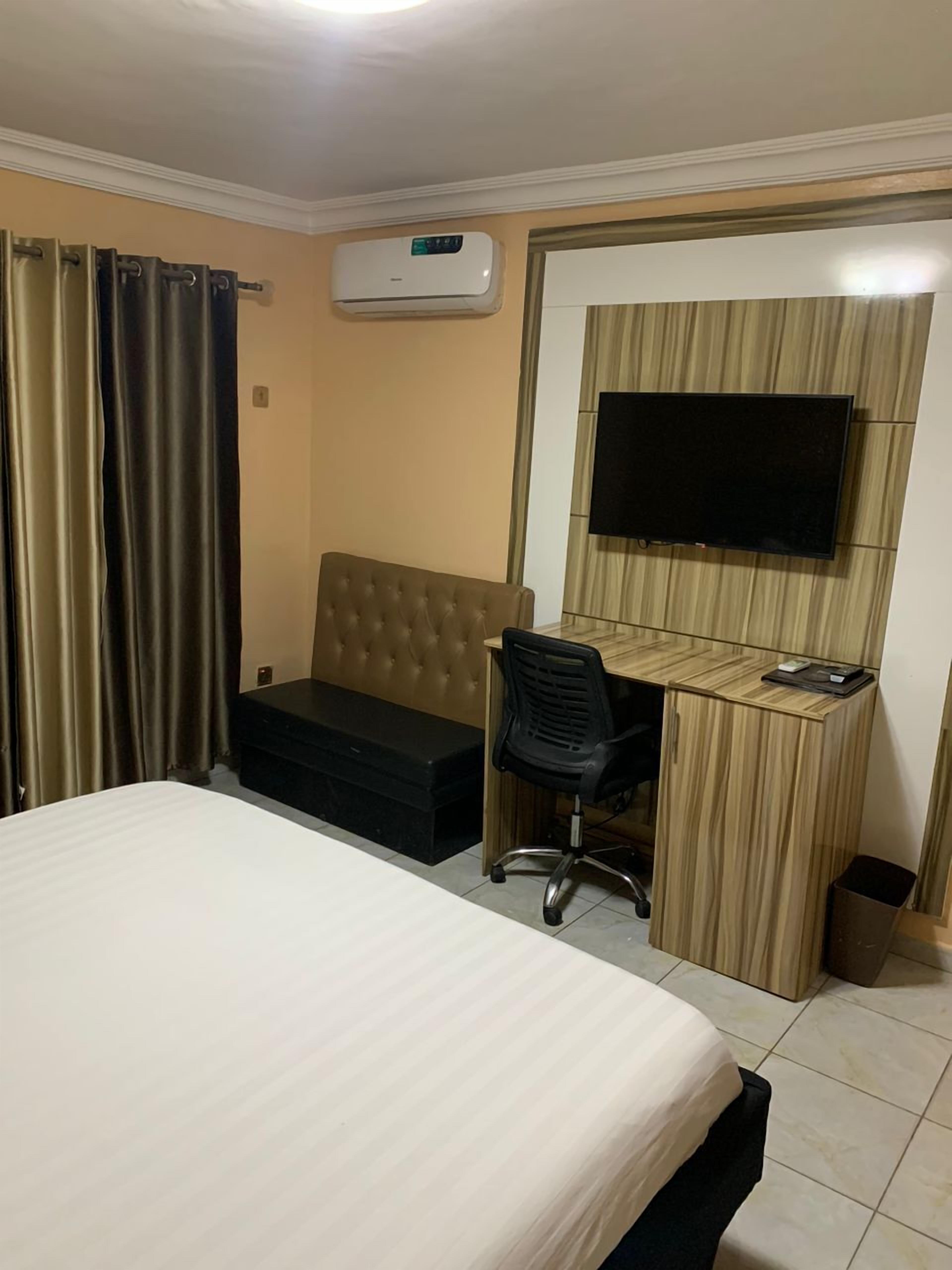 A hotel room with a bed, a desk with a chair, a television mounted on the wall, and an air conditioning unit.