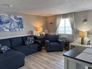 Un salón con un sofá modular, un sillón pequeño, una mesita de noche con una lámpara, un adorno de pared y una ventana con cortinas.