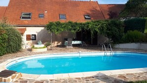A swimming pool with stone tiles, a covered patio with a table and chairs, and a building with a red roof and windows. - Domaine de la Vergnolle (Roches)