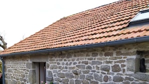 A stone building with a tiled roof, a wooden door, and a small outdoor area with a potted plant and a metal container. - Domaine de la Vergnolle (Roches)