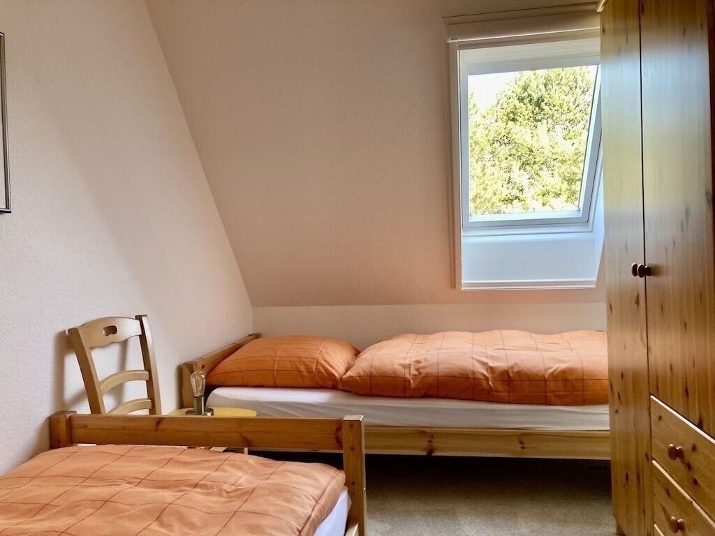A small, cozy room with a sloped ceiling, a single bed with orange bedding, a wooden chair, and a window with a view of trees.