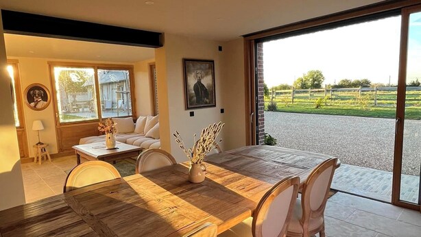 A dining area with a wooden table and chairs, a sofa, and a large window with a view of a green landscape.