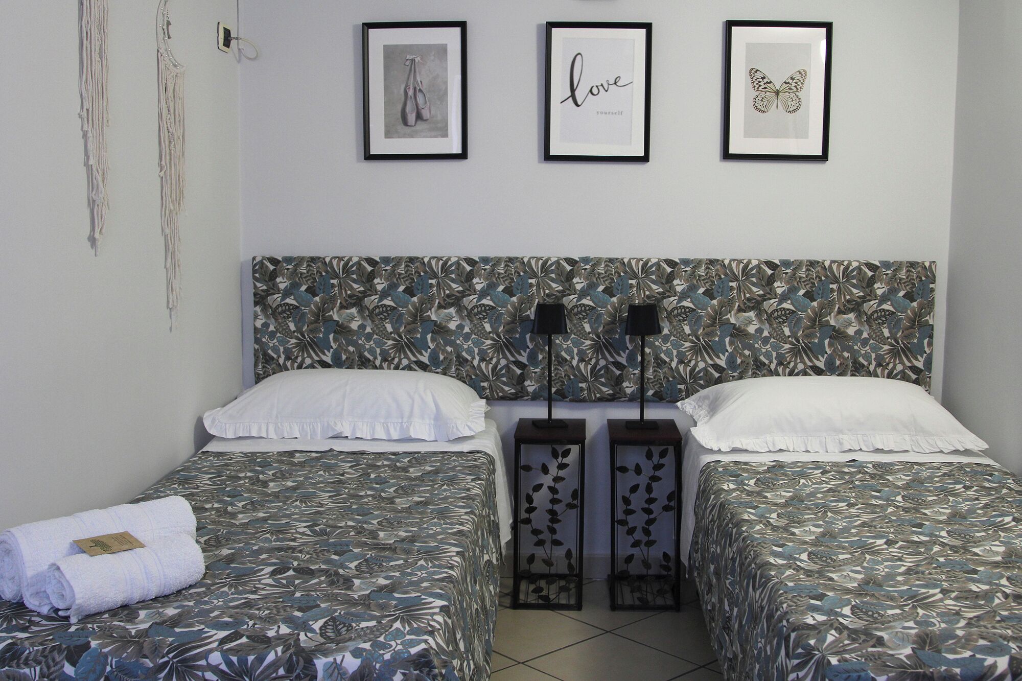 A bedroom with two beds, each with a floral-patterned bedspread, white pillows, and a headboard with a leaf design. There are three framed pictures on the wall above the beds.