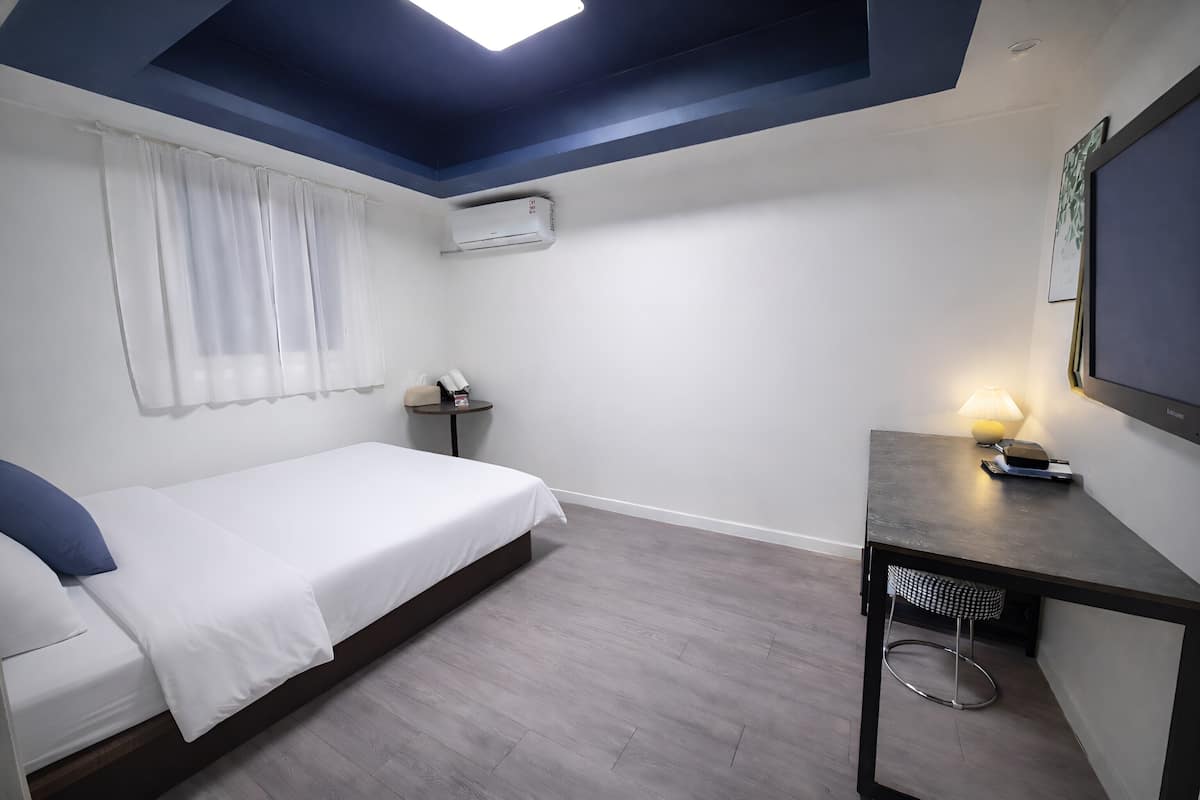 A bedroom with a bed, bedside table, lamp, air conditioning unit, and a small round table.