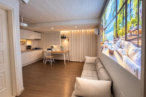 A modern living room with a wooden floor, a dining area with a table and chairs, a sofa, and a large window with a view of a beach.
