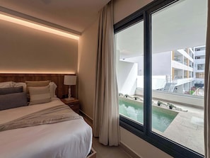 Ein Hotelzimmer mit einem großen Fenster, einem Bett mit weißer Bettwäsche, einem hölzernen Kopfteil und einem Nachttisch mit einer Lampe.