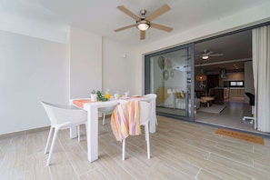 Een moderne eetruimte met een witte tafel, twee stoelen en een plafondventilator. Een glazen deur leidt naar een keuken.