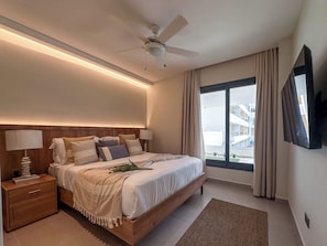 Een moderne hotelkamer met een houten bed, nachtkastjes met lampen, een plafondventilator, een groot raam met gordijnen en een flatscreen televisie aan de wand.