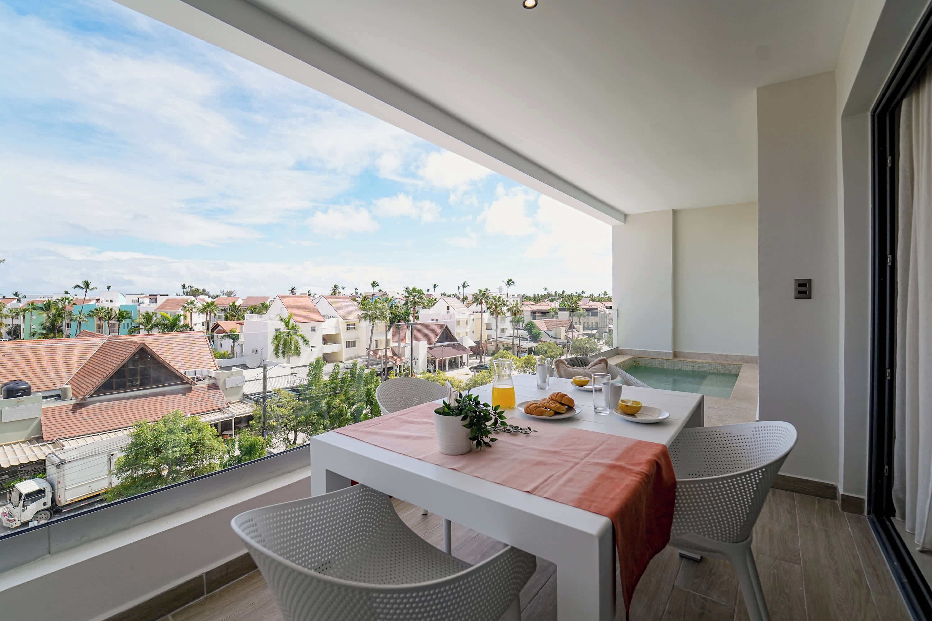 Un balcón con una mesa lista para el desayuno, con vistas a una zona residencial con casas y palmeras.