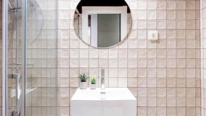 Une salle de bain avec un lavabo blanc, deux plantes en pot, un miroir rond au-dessus du lavabo et un espace douche.
