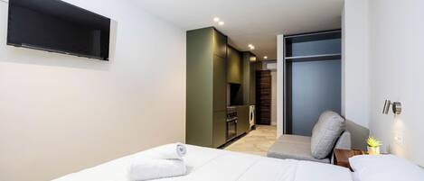 A modern hotel room with a neatly made bed, a small seating area, and a kitchenette in the background.