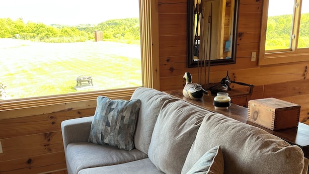 A cozy cabin interior with a beige sofa, a wooden table, and a view of a green landscape through large windows. - Charming secluded 2-bedroom cabin with WiFi, AC in peaceful Richland Center (Richland Center)