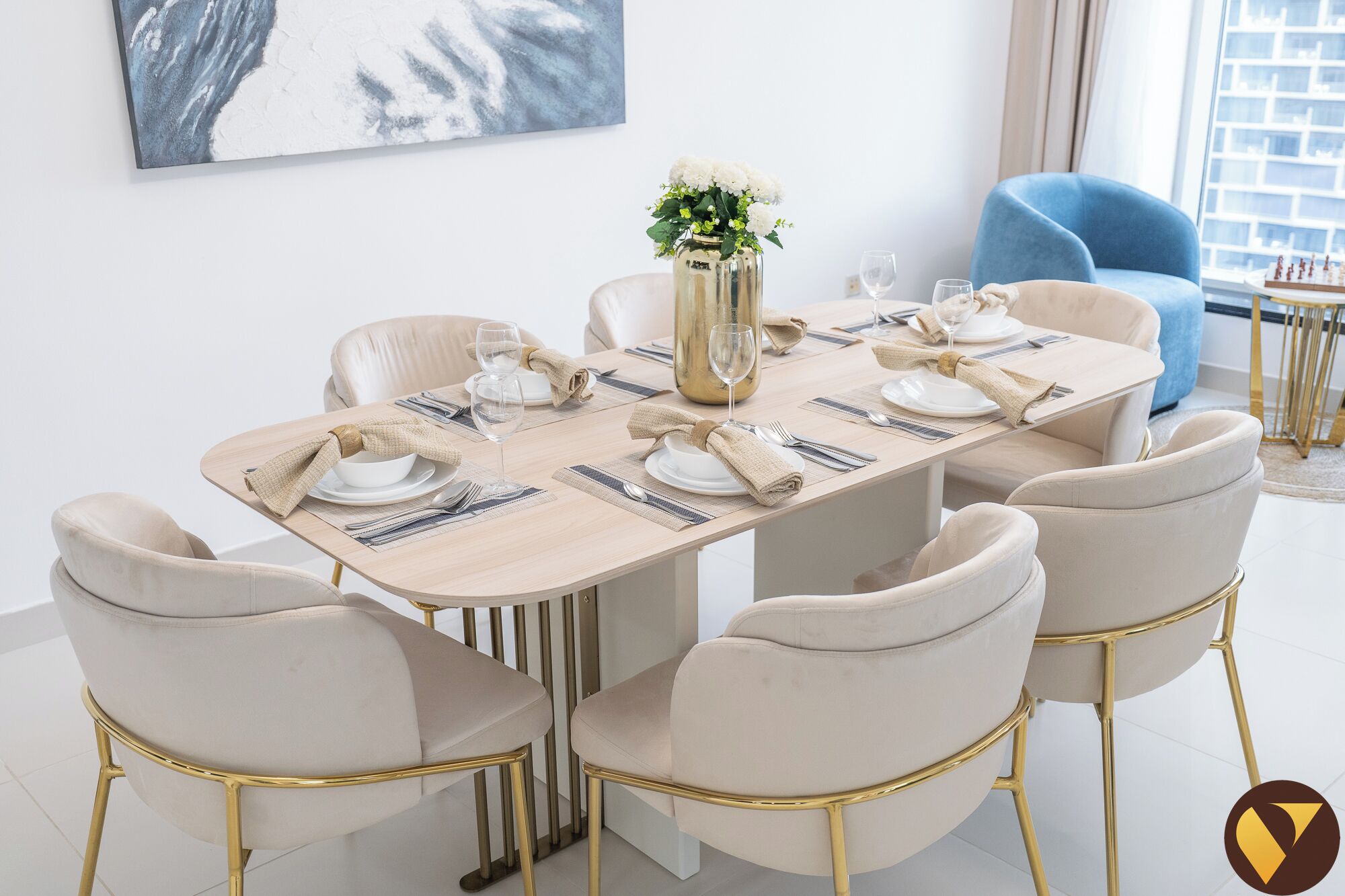 A dining area with a rectangular table set for six, surrounded by six beige upholstered chairs with gold legs. A vase with white flowers is placed on the table, and a painting is visible on the wall behind the table.