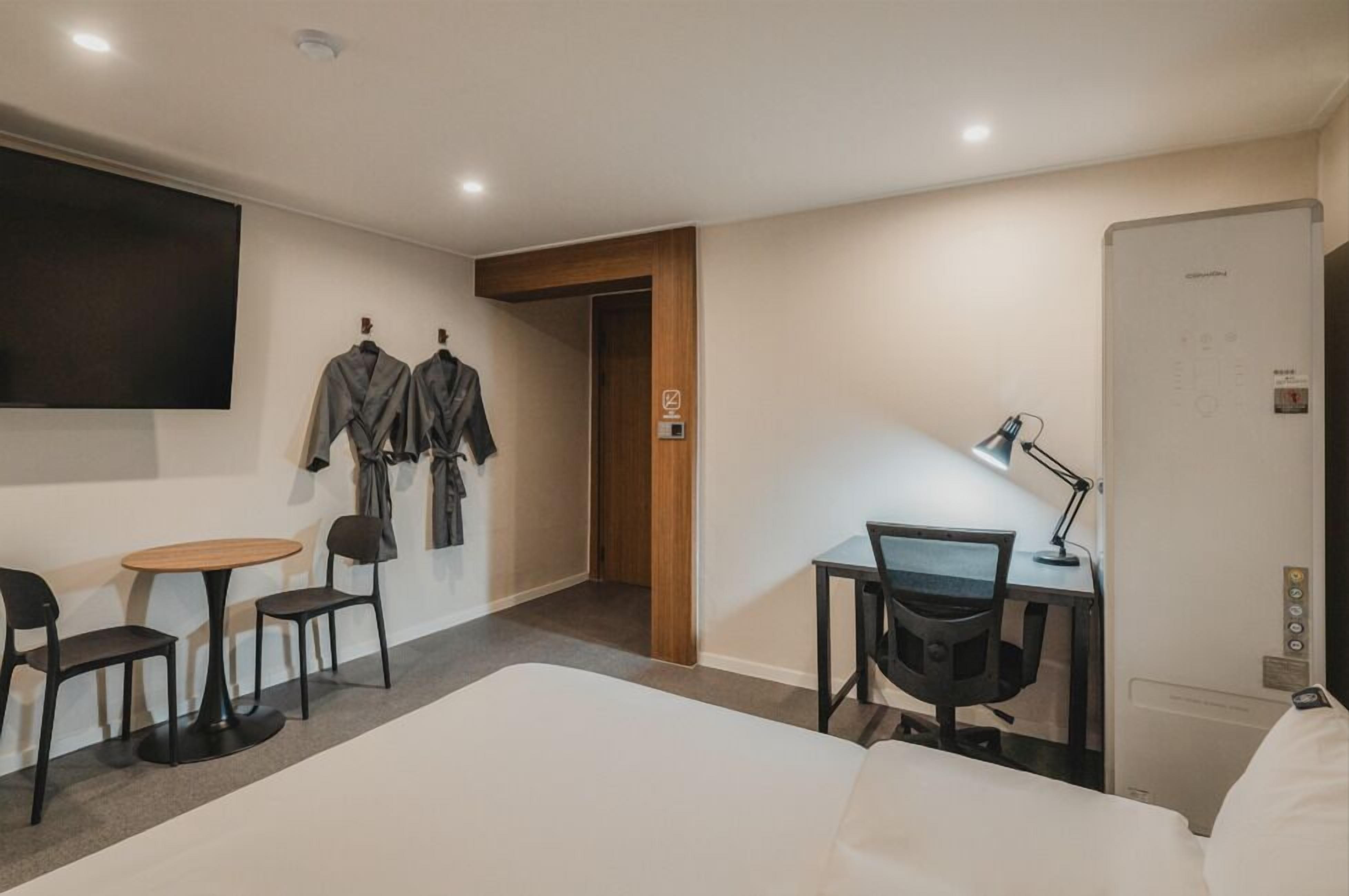 A hotel room with a bed, a desk with a chair, a small table with two chairs, a TV mounted on the wall, and a coat rack with jackets.