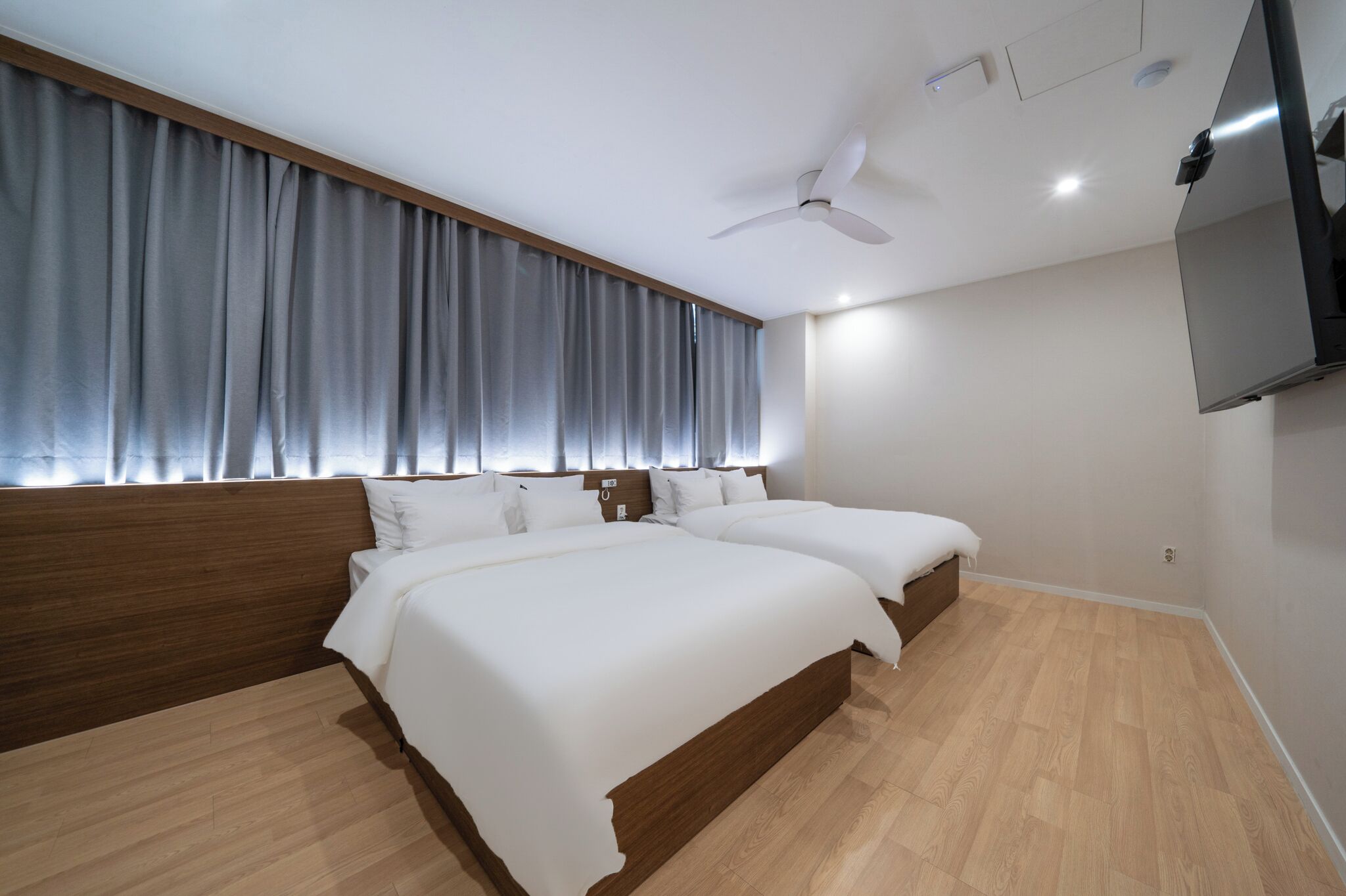 Ein modernes Hotelzimmer mit zwei Betten, einem Deckenventilator, einem Flachbildfernseher und einem großen Fenster mit Vorhängen.